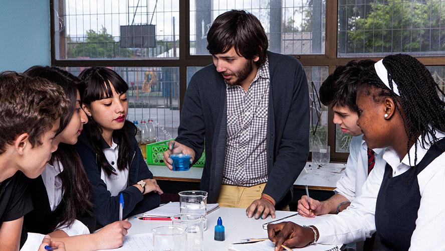 A light skinned young man with brown hair and a beard demostrates a science experiment using beekers to a diverse group of teenagers